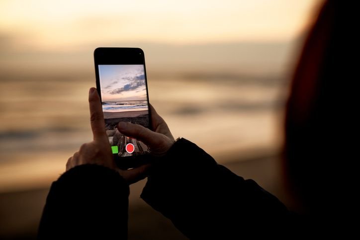 M&atilde;o feminina segurando iPhone 14 Pro com a tela ligada, fazendo um v&iacute;deo da paisagem de praia ao fundo.