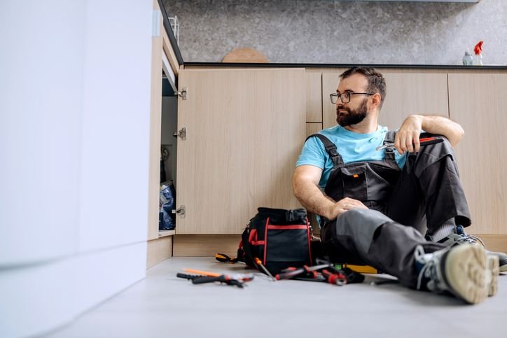 Homem sentado no ch&atilde;o da cozinha realizando manuten&ccedil;&atilde;o no arm&aacute;rio, com ferramentas espalhadas ao redor, durante o ajuste das dobradi&ccedil;as.