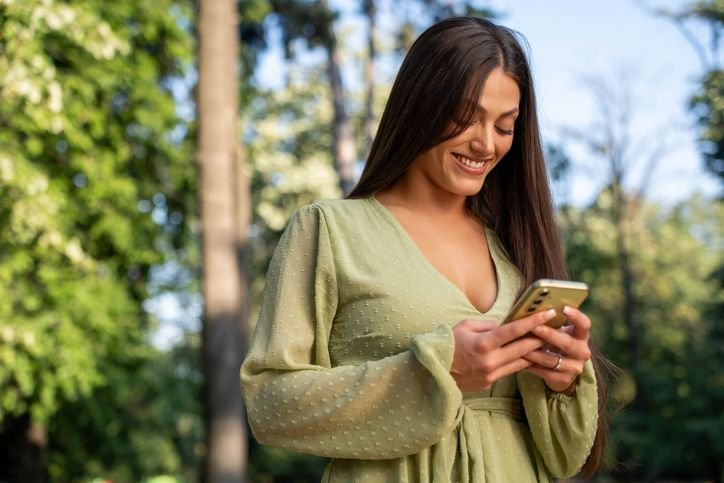 Mulher sorrindo enquanto usa smartphone em uma rua, em cena lifestyle que destaca o uso do celular na rotina.