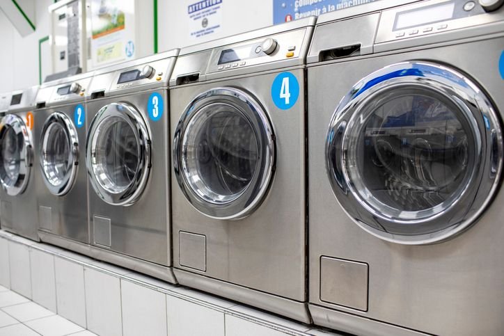M&aacute;quinas de lavar roupa inox em uma lavanderia, todas alinhadas uma do lado da outra.