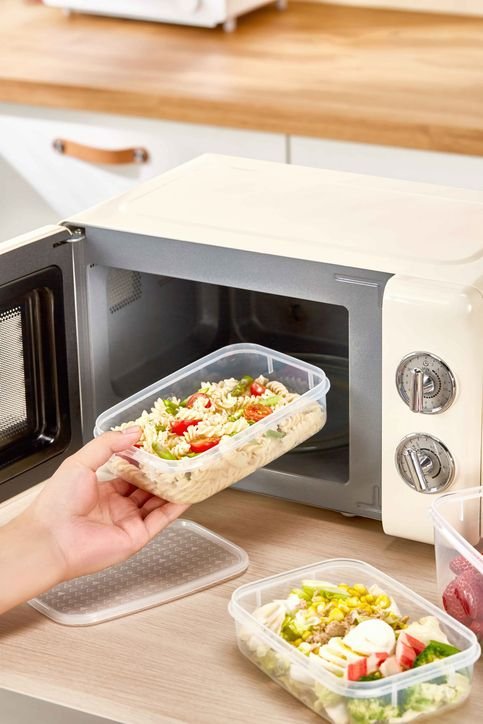 uma mão colocando um tupperware no microondas com salada e macarrão