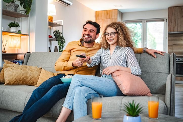 Casal sorridente no sof&aacute; de uma sala moderna, assistindo a uma mini tv digital, com suco e plantas ao lado na mesa de centro.