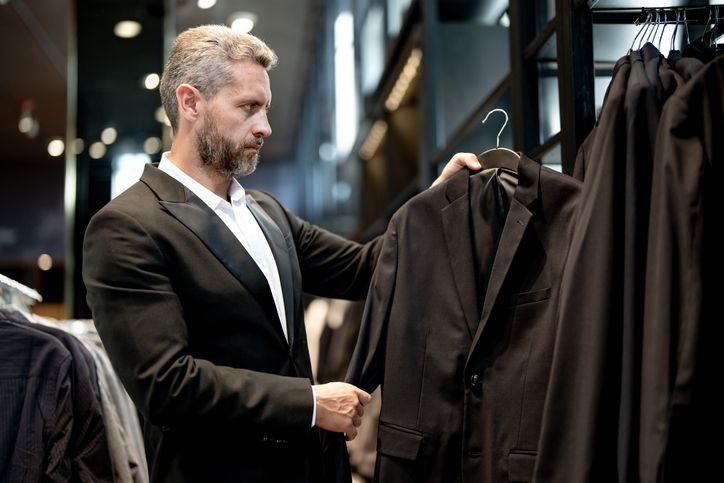 homem vestindo um terno preto está numa loja olhando outros ternos