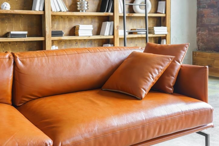 Sof&aacute; de couro marrom de estilo cl&aacute;ssico em ambiente moderno com estante e livros ao fundo.