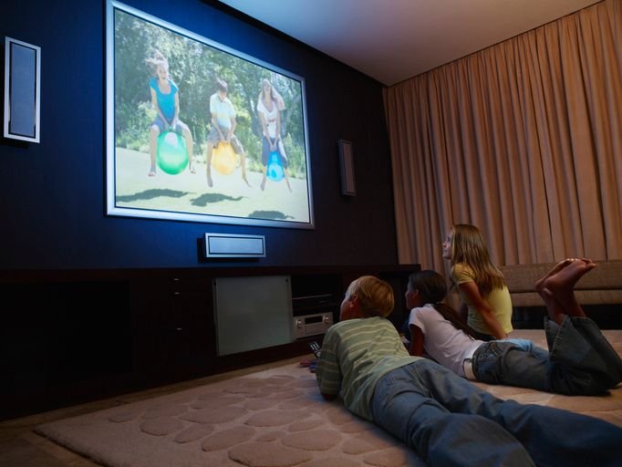 três crianças sentadas confortavelmente no tapete da sala assistindo à TV 80 polegadas 4K fixada na parede. As cortinas estão fechadas.