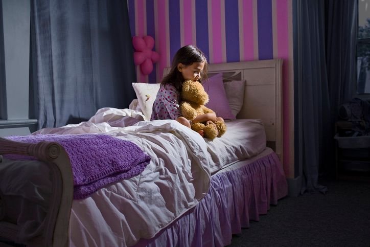 Menina sentada na cama em um quarto infantil com paredes listradas em tons de rosa e roxo, abraçando um ursinho de pelúcia., jogo de lençol solteiro branco e lilás