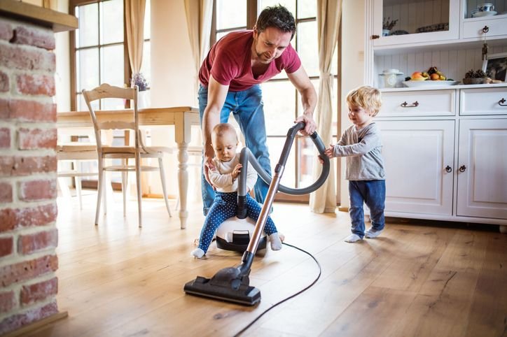 Homem com dois filhos crian&ccedil;a, usando aspirador de p&oacute; Philco no c&ocirc;modo com janelas grandes e cortinas amarelas, limpando a casa.