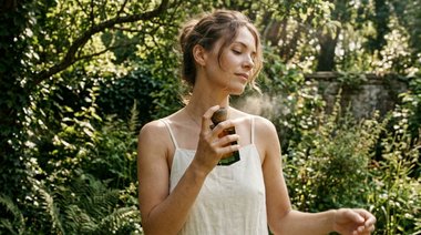 Mulher aplicando perfume amadeirado feminino importado ao ar livre em um jardim durante o verão, cercada por vegetação